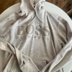 Grå hoodie från Hugo Boss - Snygg ljusgrå hoodie från Hugo Boss med stort broderat BOSS-tryck på bröstet. Tröjan har ribbade detaljer på ärmar och huva, dragsko i halsen och en klassisk känguruficka. Perfekt för en chill och stilren look.