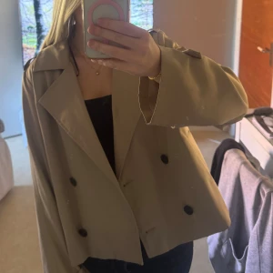  trenchcoat från Vila - Snygg croppad trenchcoat från Vila i beige med dubbelknäppning och svarta knappar. Jackan har bred krage, långa ärmar med knäppdetalj vid handlederna och en loose passform. Perfekt för vår och höst när du vill ha en stilren look.