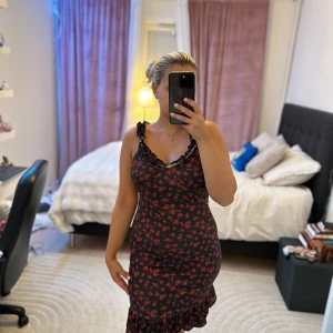 Svart blommig volangklänning från SHEIN - Supersöt svart klänning från SHEIN med röda små blommor, volangkant nertill och volangdetaljer vid axelbanden. Figurnära passform och ärmlös design, perfekt för dig som gillar romantisk stil. Materialet är mjukt och stretchigt.