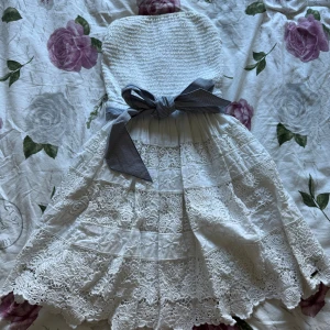 Vit spetsklänning med rosett  - Superfin vit kortklänning med smockad överdel och tunna axelband. Den är ifrån Abercrombie & Fitch. Kjolen har flera lager med spetsdetaljer och en bred, randig rosett i midjan. Perfekt för sommarens alla tillfällen! Materialet är mjukt och luftigt bomull.