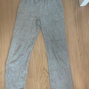 Grå mjukisbyxor Juicy Couture - Grå mjukisbyxor från Juicy Couture med broderad logga på fickan, snörning i midjan och bakfickor med knapp. Sköna och stilrena byxor i mjukt material, perfekta för chill dagar. Klassisk Juicy-look med raka ben och normal passform. Jag har snvänt de 2-3 gånget. Säljet av ingen användning och är för korta