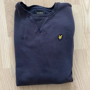 Mörkblå sweatshirt från Lyle & Scott - Snygg mörkblå sweatshirt från Lyle & Scott Junior med klassisk rund hals och gul broderad logga på bröstet. Tröjan har långa ärmar och ribbade muddar vid ärmslut och nederkant. Perfekt för dig som gillar stilrena och enkla plagg.