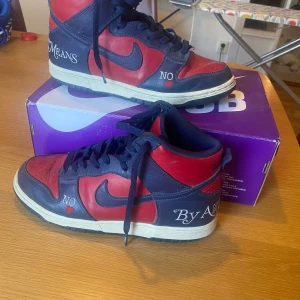 Nike Dunk High Supreme Team röd/marin - Nike Dunk High Supreme Team i rött och marinblått skinn med broderade detaljer som 'NO', 'BY ANY MEANS' och 'Supreme Team' på hälen. Klassisk siluett med snörning, vit mellansula och svart yttersula. Perfekt för dig som gillar streetwear och unika sneakers. 