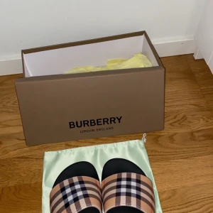 Burberry sandaler med klassiskt rutmönster - Säljer ett par Burberry sandaler med det ikoniska beige, svarta och röda rutmönstret. De har svart sula och bred rem i syntetmaterial. Perfekta för sommaren och ger en lyxig touch till din outfit.kommer med box men utan kvitto!