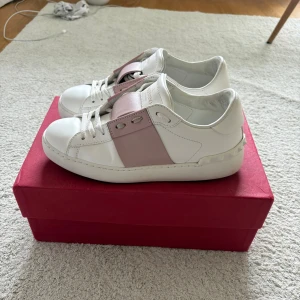 Valentino Garavani Open sneakers vit/rosa - Snygga Valentino Garavani Open sneakers i vitt skinn med breda ljusrosa detaljer över vristen. Skicket är 9/10 väldigt fina! 100% äkta. Lyxig och stilren design som lyfter din outfit. Storlek 35,5 men sitter som 36-37!