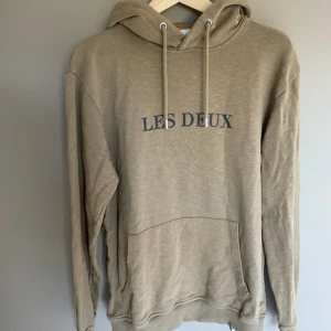 Beige hoodie från Les Deux  - Säljer en beige hoodie från Les Deux i storlek M. Tröjan har huva med snörning, stor magficka och tryck med 'LES DEUX' på bröstet. Mjuk bomullskvalitet och ribbade muddar vid ärmslut och nederkant. Perfekt för chill dagar och enkel att matcha.