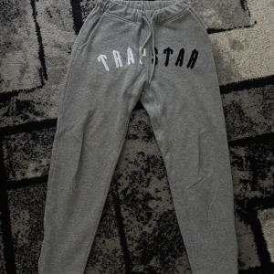 Trapstar joggers - Säljer ett par grå mjukisbyxor från Trapstar med stor logotyp i vitt och svart över framsidan. Byxorna har snörning i midjan och är tillverkade i en mjuk mix av bomull och polyester. Perfekta för chill eller streetwear och har en loose passform och är jätte bekväma. Storleken är xs men de är stora i storlek så skulle säga S.