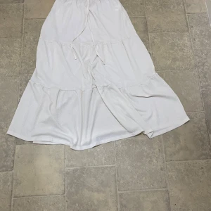 Vit långkjol med volanger och snörning - Superfin vit långkjol med volanger och snörning i midjan. Kjolen har en luftig och flowy känsla, perfekt för varma dagar. Den har flera lager och en lätt struktur i tyget som ger extra dimension till looken.