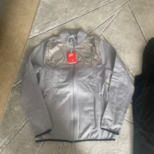 Nike silvergrå tracksuit med dragkedja  - Nike vindjacka i silvergrått med hel dragkedja och hög krage. Snygg detalj med grön broderad Swoosh på bröstet och texten 'NEVER STOP RUNNING' på ena ärmen. Jackan har släta fickor och svarta kantdetaljer. Perfekt för dig som gillar sportig stil.