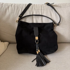 Svart axelväska från River Island (oanvänd)  - Snygg svart axelväska från River Island i mocka med detaljer i skinn och guld. Väskan har justerbar axelrem, handtag och dekorativa tofsar framtill. Magnetlås och innerfack med dragkedja gör den både praktisk och stilren.