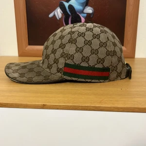 Gucci keps - Snygg keps från Gucci med klassiskt GG-mönster i beige och brunt. På sidan finns den ikoniska gröna och röda randen. Justerbar rem bak i svart skinn och insida i svart tyg. Perfekt för dig som vill ha en lyxig streetstyle-accessoar.