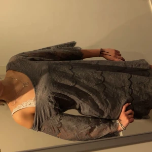 Grå spetsklänning med knappar - Säljer en grå kortklänning i spets med trekvartsärm och snygga knappar längs ryggen. Klänningen har ett vackert blommigt spetsmönster och offshoulder-design som ger en cool vibe. Perfekt för dig som gillar detaljer och vill sticka ut lite extra.