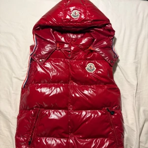 Röd dunväst med huva från Moncler - Snygg röd dunväst från Moncler med glansig finish och stor huva. Västen har dragkedjor i sidorna, tryckknappar och Moncler-logga på bröstet och huvan. Perfekt för lager-på-lager och ger en sportig vibe till din outfit. Från CN