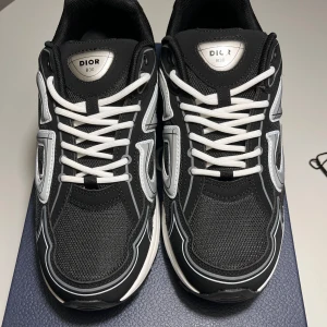 Dior B30 svarta sneakers med silverdetaljer - Dior B30 sneakers i svart mesh och syntet med vita och silverfärgade detaljer. Snygg CD-logga på sidan och bak, samt vita snören och vit sula. Sportig och modern design som sticker ut, perfekt för dig som gillar exklusiva sneakers.