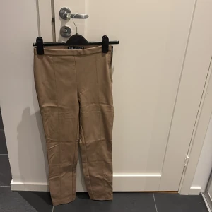 Beiga skinnbyxor från Zara, stl S - Snygga beiga byxor i skinnimitation från Zara. De har raka ben, hög midja och markerade sömmar framtill för en clean look. Perfekta att styla med en oversized hoodie eller en tight topp för en trendig vibe.