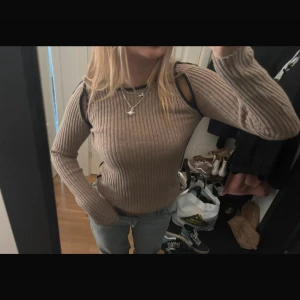 Beige ribbad tröja med cut out-detaljer - Säljer en beige ribbstickad tröja med lång ärm och coola cut out-detaljer vid axlarna. Tröjan har svarta kanter runt hals och axlar för en extra kontrast. Perfekt för dig som vill ha en unik och trendig look.