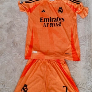 Real Madrid Vini Jr. orange matchtröja M - Säljer en orange Real Madrid fotbollströja med Vini Jr. och nummer 7 på ryggen. Adidas ClimaCool-material, kortärmad med svarta detaljer och klubbmärke på bröstet. Snyggt mönster och Emirates Fly Better-tryck på framsidan. Perfekt för fans av Vini Jr. och Real.