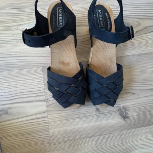 Svarta Moheda träskosandaler - Snygga svarta sandaler från Moheda med flätad ovandel i skinn och klassisk träbotten. Skorna har justerbart spänne runt ankeln och en låg klack. Perfekta för sommarens alla tillfällen och ger en cool retrovibe.