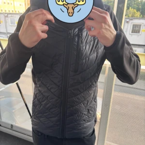 Svart jacka från McKinley - Svart hybridjacka från McKinley med mjuka ärmar och huva. Jackan har dragkedja framtill och en bröstficka med dragkedja. Perfekt för lager-på-lager och chill höstväder.