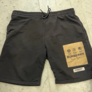 Svarta Burberry shorts med patch - Svarta shorts från Burberry med beige patch och logga på ena benet. De har elastisk midja med snörning och är tillverkade i mjuk bomull. Perfekta för en chill och stilren look.