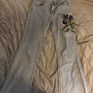 Ed Hardy ljusgrå jeans med brodyr - Säljer ett par ljusgrå jeans från Ed Hardy med snygg brodyr på ena benet. Jeansen har utsvängda ben och klassisk femficksdesign. Materialet är jeans och de har en cool streetstil med broderad logga på baksidan.