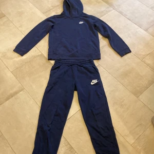 Mörkblå Nike hoodie & mjukisbyxor - Hej! Säljer ett Nikeset som inkluderar hoodie i mörkblått med klassisk logga på bröstet och mjukisbyxor. Tröjan har luva, långa ärmar och en stor magficka framtill. Byxorna har muddar vid bensluten och resor vid midjan. Båda har bomull inuti för värma och komfort. Finns även att köpa enskilt. Mjukt och skönt, perfekt för chill och mysiga höstdagar! Tveka inte att höra av dig vid frågor!