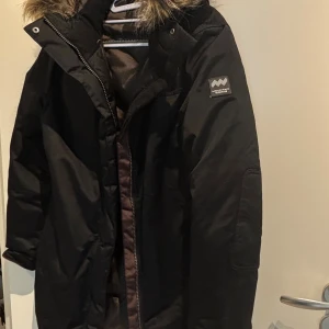 Svart dunjacka från Mountain Works - Svart dunjacka från Mountain Works med huva och snygg fuskpäls. Jackan har dragkedja framtill, långa ärmar och flera praktiska fickor. Insidan är quiltad och jackan har en normal passform. Perfekt för kalla vinterdagar. Lite slitage på insidan, men inget som påverkar funktion.