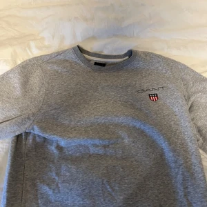Grå sweatshirt från GANT i storlek L - Snygg grå sweatshirt från GANT med broderad logga och emblem på bröstet. Tröjan har rund hals, långa ärmar och ribbade muddar vid hals, ärmslut och nederkant. Perfekt för chill dagar och enkel att matcha med jeans eller joggers. Storlek L men passar även M, knappt använd, nypris 899kr, priset kan diskuteras.