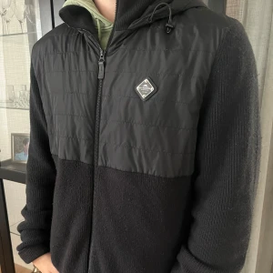 Svart hybridjacka från J.Lindeberg - Svart cardigan från J.Lindeberg med quiltad framsida i syntet och stickade ärmar och baksida. Cardiganen är i storlek XL men passar dig som har L i vanliga fall. Den är i riktigt bra skick och riktigt snygg nu till hösten! Bara att höra av sig vid någon fundering!😃