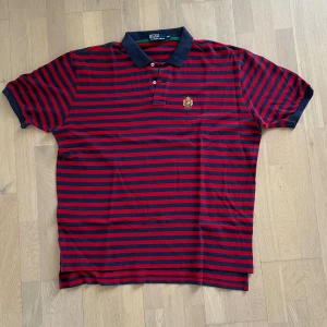 Randig pikétröja från Polo Ralph Lauren - Snygg pikétröja från Polo Ralph Lauren i rött och marinblått med horisontella ränder. Klassisk krage, knäppning med två knappar och broderad logga på bröstet. Perfekt för en stilren och sportig look.