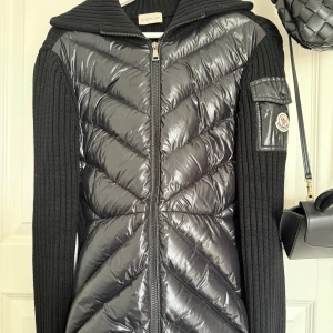 Svart cardigan från Moncler - Svart cardigan från Moncler med glansig quiltad front och ärmficka med logga. Jackan har ribbstickade ärmar och rygg, dragkedja framtill. Två stängbara fickor, orginalprislapp finns kvar!