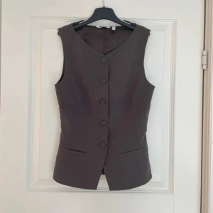 Brun väst från Weekday med knappar - Snygg grå väst från Weekday i klassisk modell med v-ringning och fem knappar framtill. Västen har två fejkade fickor och är figursydd för en slim passform. Perfekt att styla över en t-shirt eller skjorta för en cool look.