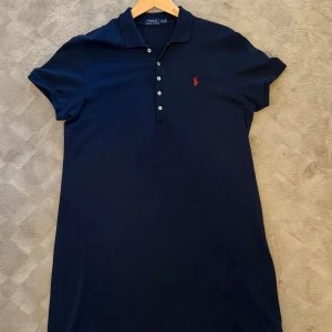 Säljer Marinblå pikéklänning från Polo Ralph Lauren! - Klänningen är som helt ny och endast använd ett fåtal gånger.  Storlek: XL Snygg marinblå kortklänning från Polo Ralph Lauren med klassisk pikékrage och knappslå framtill. Klänningen har korta ärmar och det ikoniska röda Polo-logot broderat på bröstet. 