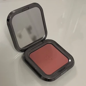KIKO kompakt rouge  - Snyggt kompakt rouge från KIKO i en rosad nyans, perfekt för att ge kinderna en fräsch färg. Kommer i en stilren, kvadratisk dosa med spegel och metallisk finish. Smidig att ha med i väskan och enkel att applicera med borste. 