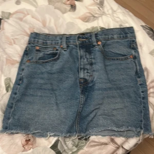 Blå jeanskjol med rå kant - Säljer en klassisk blå jeanskjol från 157 med rå, fransig kant nertill. Kjolen har fem fickor, bälteshällor och dragkedja fram. Perfekt för dig som gillar en avslappnad och trendig stil.