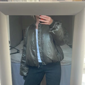 Olivgrön puffer bomberjacka Samsøe - Olivgrön puffer bomberjacka från Samsøe Samsøe med Thermore-foder av återvunna fibrer. Jackan har ribbad krage, långa ärmar, två fickor fram och dragkedja. Oversized passform och glansigt material ger en cool vibe.  L men liten i storlek