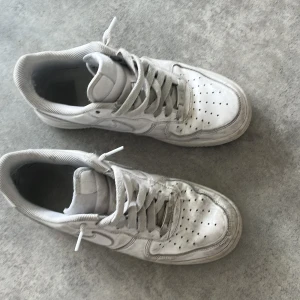 Nike Air Force 1 vita sneakers - Klassiska Nike Air Force 1 sneakers i helvitt skinn med perforerad tå, rund tå och platt sula. Snygg low-top modell med snörning och ikonisk Swoosh-logga på sidan. Perfekta för dig som gillar streetstyle och tidlös design.