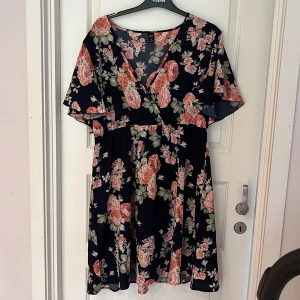 Blommig kortklänning från SHEIN Curve - Supersöt kortklänning från SHEIN Curve med marinblå botten och stora rosa blommor. Klänningen har v-ringning, korta vida ärmar och markerad midja. Perfekt för dig som gillar flowy och romantisk stil. Materialet är lätt och luftigt.