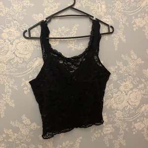 Svart spetsig linnetopp med rosett - Säljer en svart ärmlös linnetopp i spets med v-ringning och liten rosett framtill. Toppen är genombruten med blommigt spetsmönster och har en feminin, croppad passform. Perfekt att styla med jeans eller kjol för en trendig look.
