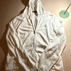 Vit hoodie från Polo Ralph Lauren - Vit hoodie från Polo Ralph Lauren med dragkedja och huva. Klassisk broderad logga i marinblått på bröstet. Tröjan har långa ärmar och fickor framtill. Tillverkad i mjuk bomull för skön känsla. Perfekt för chill dagar och enkel att matcha.