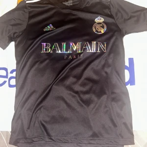 Svart Real Madrid x Balmain tröja - Snygg svart fotbollströja från Adidas med Real Madrid-emblem och Balmain Paris-logga i holografiskt tryck på bröstet. Klassisk passform med korta ärmar och rund hals. Perfekt för dig som vill sticka ut på planen eller gatan.