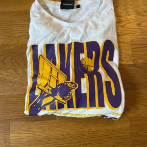 Vit Lakers t-shirt från New Era, M - Vit t-shirt från New Era med stor Los Angeles Lakers-print i lila och gult på framsidan. Klassisk passform med rund hals och korta ärmar. Perfekt för dig som älskar basket och vill visa ditt Lakers-stöd. Materialet är mjuk bomull.