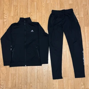 Svart Adidas träningsset med dragkedja - Snyggt svart träningsset från Adidas med jacka och byxor. Jackan har hög krage, hel dragkedja och två fickor med dragkedja. Byxorna har resår och snörning i midjan samt Adidas-logga i silver på ärm och ben. Perfekt för gym eller sport.