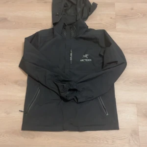 Svart vindjacka från Arc'teryx M - Svart vindjacka från Arc'teryx i storlek M. Jackan har huva, dragkedja framtill och två snedställda fickor med dragkedja. Snygg minimalistisk design med Arc'teryx-logga på bröstet och diskreta detaljer. Perfekt för blåsiga dagar och outdoor.