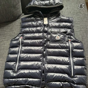 Svart dunväst från Moncler med huva - Snygg svart dunväst från Moncler med avtagbar huva, två dragkedjefickor och en bröstficka med logga. Västen har quiltad design och blanka detaljer som ger en exklusiv känsla. Perfekt för lager-på-lager och streetstyle.