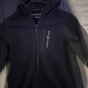 Sail racing  - Säljer min sail racing hoodie eftersom den är för liten för mig. Hoodien har inget fel i sig det enda som är ett ”problem” är att det har försvunnit lite tyg på sidan av ärmen som jag visar i bild ”3” men det är inget man tänker på i verkligheten 