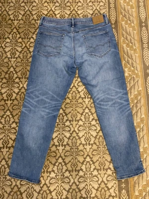 Snygga jeans från ameriacn eagle - Säljer ett par klassiska blå jeans från American Eagle med rak passform och fem fickor. Jeansen har snygga slitningar och ljusare partier framtill för en trendig look. Tillverkade i slitstark denim med läderpatch bak i midjan. Perfekta för en avslappnad stil. 