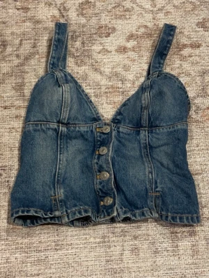 Jeanstopp från Zara - Säljer en snygg denim topp från Zara i blå denim med breda axelband och v-ringning. Toppen har dekorativa sömmar och knappar framtill som ger en cool vintagekänsla. Perfekt att styla med höga jeans eller kjol för en trendig look.