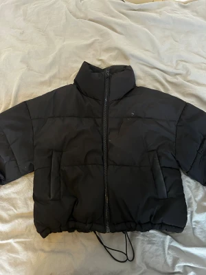 Svart croppad pufferjacka - Snygg svart pufferjacka med croppad passform och hög krage. Jackan har dragkedja framtill, två stora fickor och justerbar snörning i nederkant. Perfekt för kalla dagar när du vill ha en trendig look.