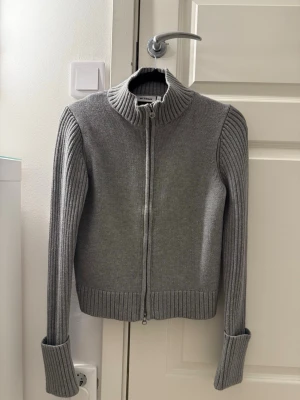 Grå ribbad ziptröja med hög krage - Snygg grå ziptröja med hög krage och ribbade detaljer på ärmar och nederkant. Tröjan har långa ärmar med breda muddar och dragkedja framtill. Perfekt för lager på lager och enkel att styla till olika outfits.